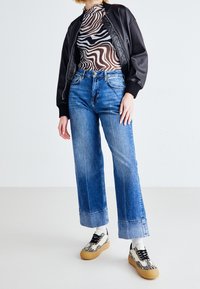 Osoba v černé kožené bundě, průhledném topu se zebřími pruhy, širokých modrých džínech s ohrnutými nohavicemi a bílých teniskách s černými pruhy.