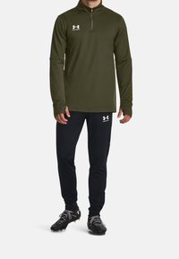 Olivgrünes Langarmshirt mit Viertel-Reißverschluss-Design, kombiniert mit schwarzer Hose und schwarzen Fußballschuhen mit weißen Akzenten.