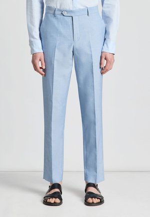 Personne vêtue d'un pantalon bleu clair sur mesure et de sandales noires ouvertes à l'avant, debout devant un fond clair uni.
