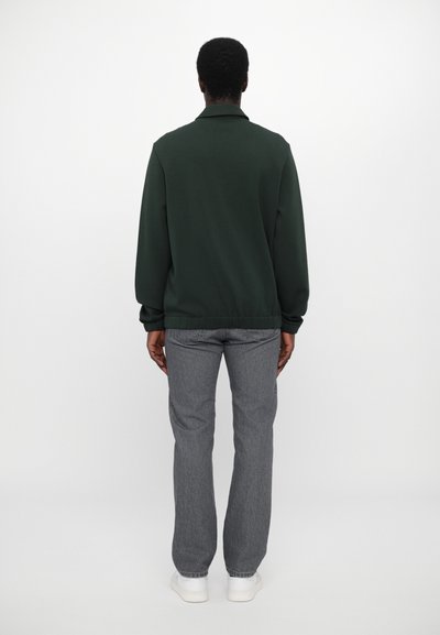 Grønn sweatshirt, lange ermer, ribbestrikkede mansjetter og nederkant, avslappet passform. Kombinert med grå jeans og hvite joggesko, sett fra baksiden.