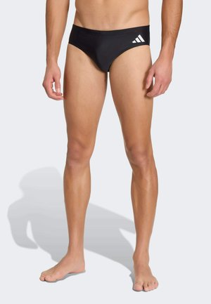 Male model wearing black swim briefs with white logo on left hip, standing barefoot on flat light surface.