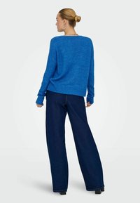 Femme aux cheveux blonds attachés en chignon, portant des boucles d'oreilles créoles, un pull bleu vif et un jean large sombre, debout, dos face à l'avant.