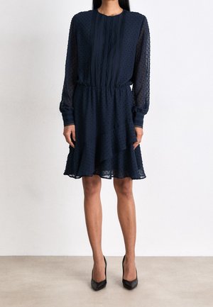 Kobieta ubrana w granatową, długą do kolan sukienkę z przezroczystej tkaniny w kropki oraz czarne szpilki z migdałowymi noskami, stojąca na gładkiej podłodze.