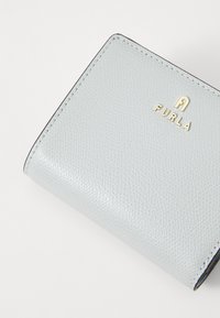 Hellgraue Lederbrieftasche mit strukturierter Oberfläche, goldfarbener "FURLA"-Logo und kontrastierenden Nähten entlang der Kanten. Kompakte rechteckige Form.