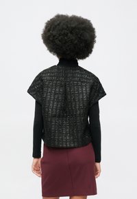 Osoba s afro frizurom nosi crni teksturirani kratki rukav top preko crnih dugih rukava i bordo suknju, gledana odostraga.