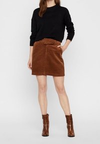 Bruine corduroy rok met een ceintuur in de taille, gecombineerd met een zwarte trui en bruine enkelboots. De stof heeft een gestructureerde uitstraling.