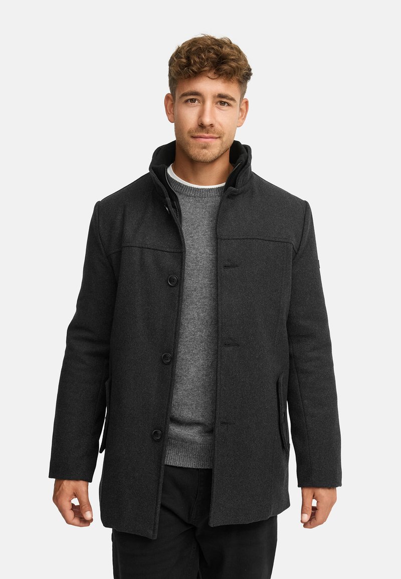 Cappotto grigio scuro in misto lana con colletto alto, chiusura a bottoni, tasche laterali e silhouette strutturata, indossato sopra un maglione grigio.