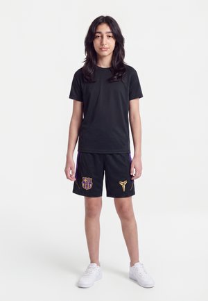 Jeune personne portant une tenue de sport noire avec les logos du FC Barcelone et de Kobe Bryant, debout, les bras détendus, sur un fond blanc uni.