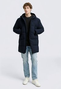 Chaqueta acolchada azul marino con capucha, con cremallera frontal, dos grandes bolsillos delanteros y textura acolchada. Llevada sobre un suéter y vaqueros.