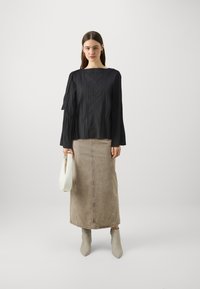 Schwarzes, strukturiertes Oberteil mit weiten Ärmeln, kombiniert mit einem beigefarbenen, knielangen Rock. Das Model hält eine weiße Handtasche und trägt graue Stiefeletten.