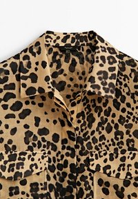 Chemise beige avec un motif imprimé léopard noir et marron foncé, col étalé, fermeture à boutons et deux poches poitrine à rabat.
