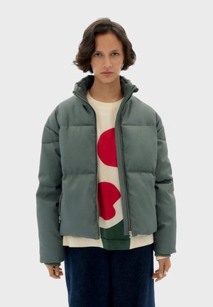 Padded, zip-up jacket in muted green, with a high collar, featuring a smooth texture and minimal quilting design. Worn over a cream shirt with red and green patterns.