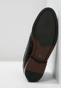 Zapato de cuero negro con diseño de brogue, mostrando una suela texturizada y un tacón marrón en contraste. Talla marcada como 37.