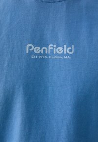Mėlynas medvilnės marškinėliai su apvalia iškirpte, baltu tekstu "Penfield" ir "Est 1975, Hudson, MA." Minkšta tekstūra, paprastas dizainas.