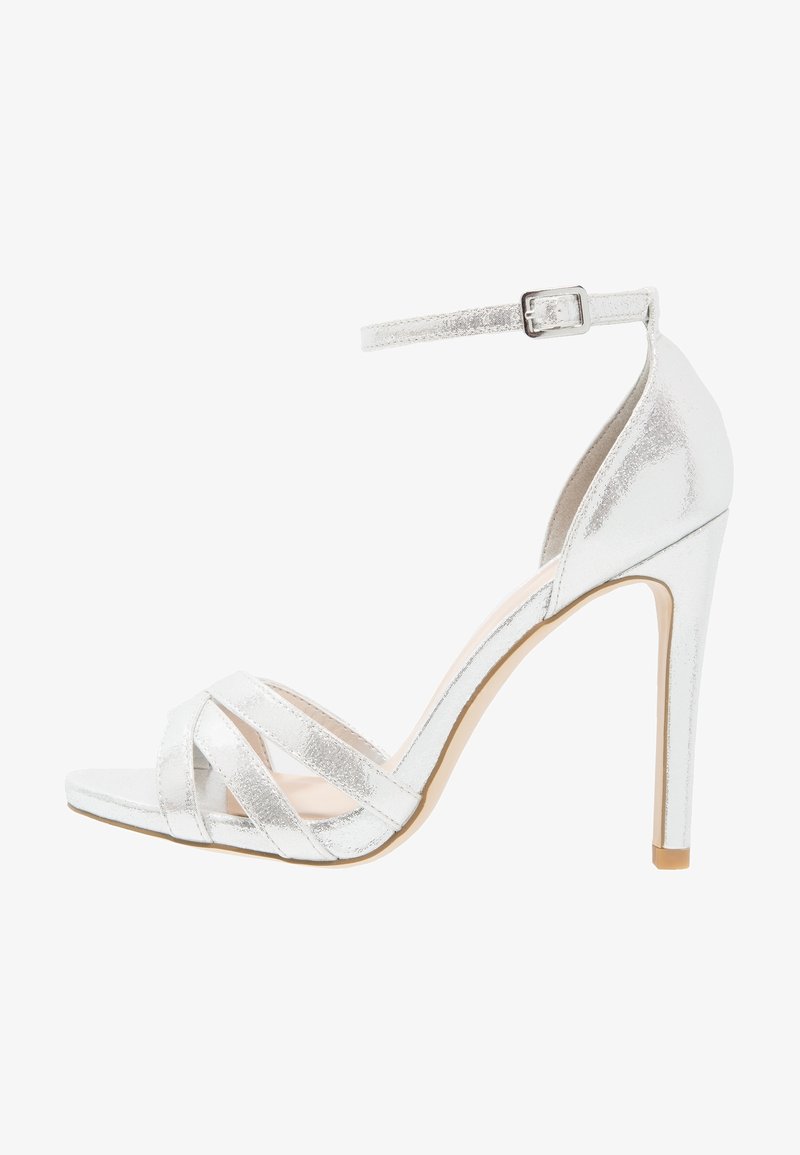 Sandale argentée à talon haut avec bride à la cheville, présentant des brides croisées à l'avant et une finition métallique lisse. Design à talon aiguille.