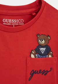 Punainen luomupuuvillainen T-paita, jossa on taskupainatus sinisessä villapaidassa olevan karhun kuvasta ja "guess" navy-värillä brodeerattuna.