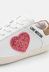 Vita lädersneakers med en rosa glittrande hjärtapatch på sidan och texten "LOVE MOSCHINO". Ljusbeige gummisula med snörningsdesign.