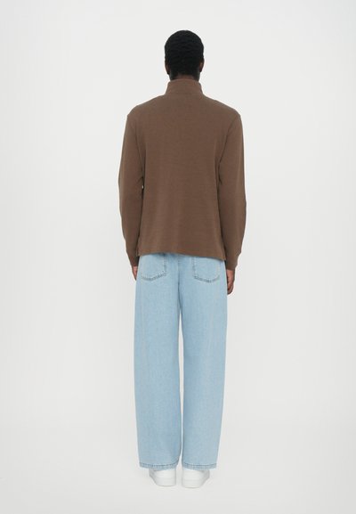 Langsleeve bruine shirt met een kraag, gecombineerd met lichtblauwe baggy jeans en witte sneakers, weergegeven vanuit de achterkant tegen een neutrale achtergrond.