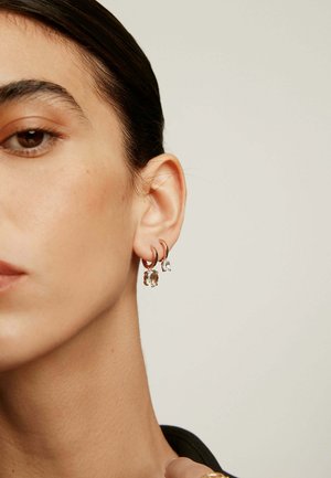 Pendientes de aro en oro rosa con piedras preciosas transparentes, que cuentan con tres colgantes distintos, uno más grande y dos más pequeños, que reflejan la luz sobre la piel.