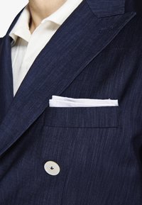 Persona vistiendo una chaqueta de traje azul marino con una camisa blanca y un pañuelo blanco en el bolsillo del pecho.