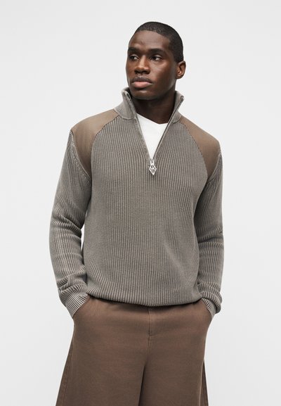 Άνδρας που φοράει πλεκτό πουλόβερ μισής ζώνης σε χρώμα ταουπέ με σουέτ επιθέματα στους ώμους και καφέ παντελόνι, στέκεται με τα χέρια στις τσέπες, κοιτώντας προς τα δεξιά.