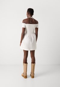 Žena s krátkými vlasy, oblečená v bílé šatech bez ramínka a béžových černých botách po kotníky, stojí čelem k hladké bílé zdi.