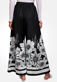 Los pantalones anchos negros presentan un diseño floral en blanco, con acentos de hojas y un dobladillo a rayas en contraste, hechos de un material suave y fluido.