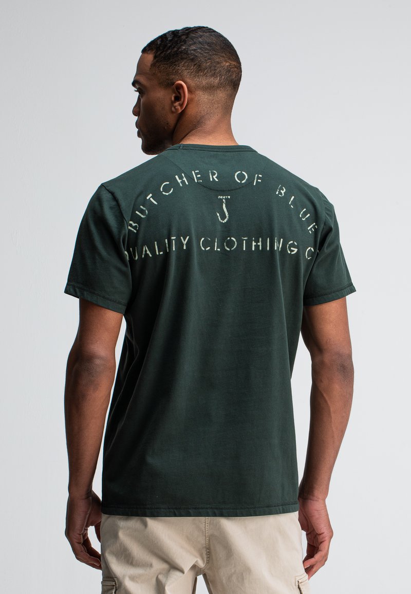 Man in een donkergroene T-shirt met vervaagde witte tekst "Butcher of Blue Quality Clothing Co" op de achterkant, gecombineerd met beige broek, staande tegen een witte achtergrond.