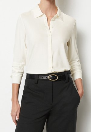 Femme portant une chemise à manches longues crème à boutons, rentrée dans un pantalon noir, avec une ceinture noire ornée d'une boucle en or ovale.