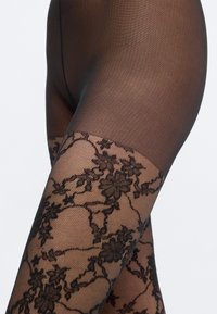 Μαύρες καλσόν με φλοράλ δαντελένιο σχέδιο στον μηρό, με διαφανή υφή και λεία επιφάνεια, δημιουργώντας οπτική αντίθεση στο σχέδιο.