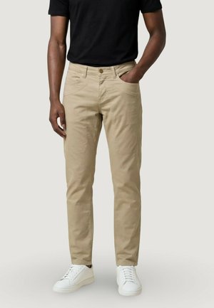 Uomo con pantaloni beige aderenti, camicia nera a maniche corte e scarpe da ginnastica bianche, in piedi con una mano in tasca contro uno sfondo semplice.