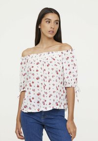 Blouse blanche à épaules dénudées avec un motif floral rouge et bleu, encolure froncée et manches courtes bouffantes. Tissu léger avec un design fluide.