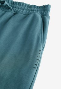 Teal sweatpants lavet af blødt stof med en elastisk talje, der har en sidelomme og "ACTIVE" grafik trykt på benet.