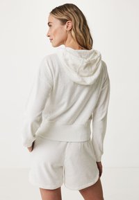 Felpa bianca in tessuto testurizzato con cappuccio regolabile e bordo a costine, abbinata a pantaloni corti bianchi coordinati con un design morbido e informale.