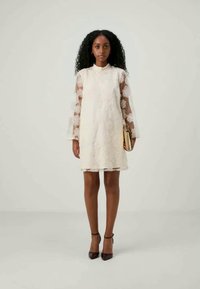 Vestido de renda em off-white com mangas longas transparentes e padrão floral. Silhueta curta, decote redondo, complementado por uma clutch e sapatos de salto.