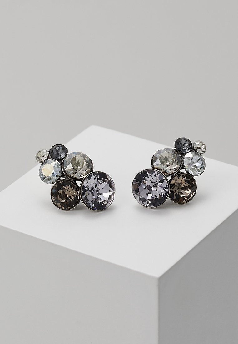 Groupe de clous d’oreilles ronds, de tailles variées, en pierres précieuses facettées grises, noires et transparentes sur un bloc d’exposition blanc.