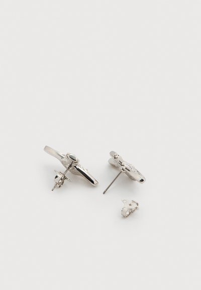 Ασημένια σκουλαρίκια μικρά με υφή στην επιφάνεια· το ένα διαθέτει κεντρική πέτρα και λεπτές διακοσμήσεις, ενώ το άλλο είναι ένα απλό κυκλικό σχέδιο.