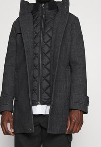 Manteau en mélange de laine gris foncé avec une doublure intérieure matelassée noire zippée, des boutons en accent, de grandes poches et un col montant. Texture douce.