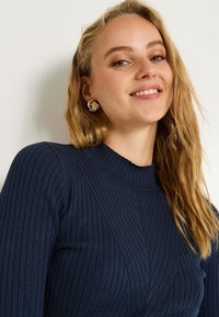 Pull à côtes navy avec un col montant et des détails angulaires, associé à des boucles d'oreilles texturées dorées. Tissu doux, coupe ajustée.