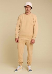 Un sweat à capuche beige et un pantalon de jogging en matière douce avec des poignets côtelés, associés à des baskets légères avec des accents gris et pastel.
