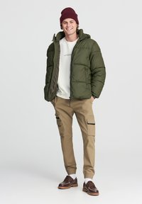 Zielona kurtka puchowa z zamkiem, w połączeniu z beżowymi spodniami cargo z bocznymi kieszeniami i prążkowanym wykończeniem oraz brązowymi butami na sznurówki.