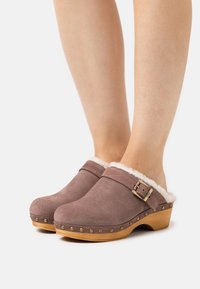 Braune Wildleder-Clogs mit einem weichen weißen Fellfutter, Holzsohle und dekorativen goldenen Nieten, ausgestattet mit einem Schnallenriemendetail.