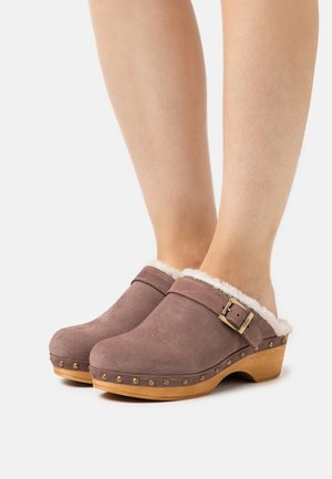 Brown suede clogs with a plush white fur lining, wooden sole, and decorative gold studs, featuring a buckle strap detail.