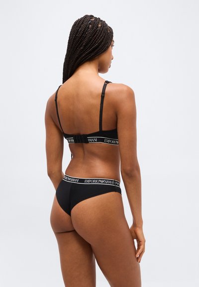 Γυναίκα με πλεγμένα μαλλιά που φοράει μαύρο σουτιέν και σλιπ Emporio Armani, εμφανιζόμενη από πίσω σε απλό ανοιχτό φόντο.