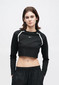 Νεαρή γυναίκα με κοντά καστανά μαλλιά, φορώντας μία μαύρη κοντή μπλούζα με μακριά μανίκια της Nike και μαύρο παντελόνι, που στέκεται μπροστά σε λευκό φόντο.