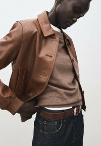 Giacca corta in pelle marrone con colletto, chiusura a bottoni e polsini a coste, indossata sopra un maglione beige e jeans in denim scuro.