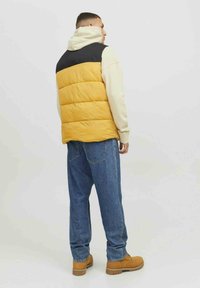 Chaleco acolchado con una parte superior negra, cuerpo amarillo y mangas beige, combinado con jeans azules y botas color canela, visto desde la parte trasera. Diseño limpio.