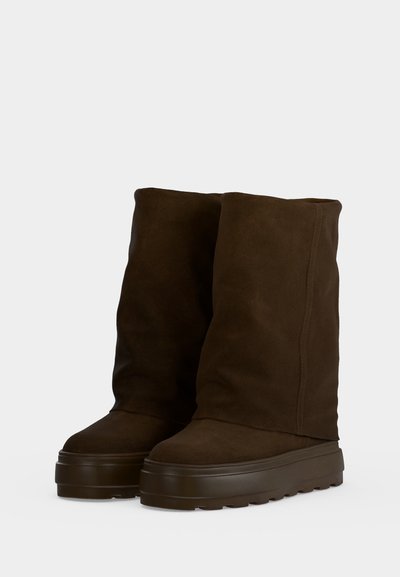Paire de bottes en daim marron foncé mi-mollet avec semelles épaisses en caoutchouc sur fond gris clair.