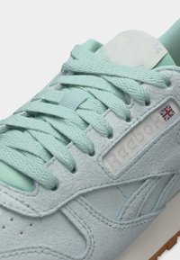 Ανοιχτό μπλε σουέντ αθλητικό παπούτσι με υφασμένους κορδόνια, λογότυπο Reebok και μια μικρή σημαία του Ηνωμένου Βασιλείου στο πλευρικό πάνελ.