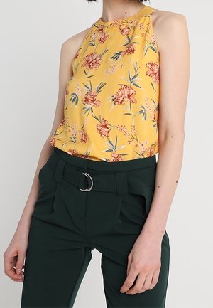 Top a balconcino giallo con fiori rossi e foglie verdi; abbinato a pantaloni a vita alta verde scuro con dettaglio in metallo argentato.